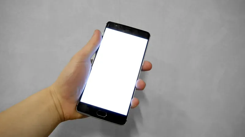 An adult hand holds a smartphone with a blank white screen against a gray wall.